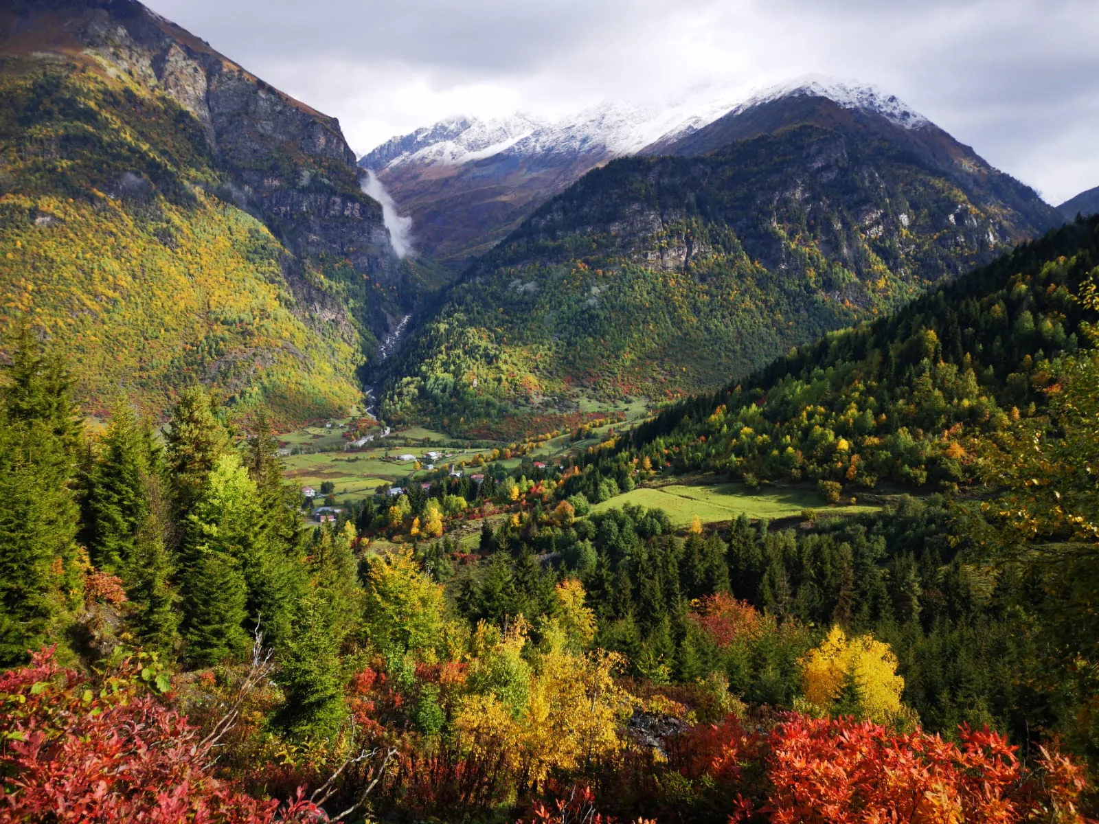 Autumn forest and a valley village below snowy peaks in Svaneti